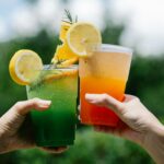 pexels-photo-8679436-8679436 Two hands holding colorful citrus lemonades in a vibrant summer outdoor setting.
